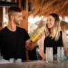 Bartender smiling while holding a shaker next to a laughing woman at a tropical outdoor bar.