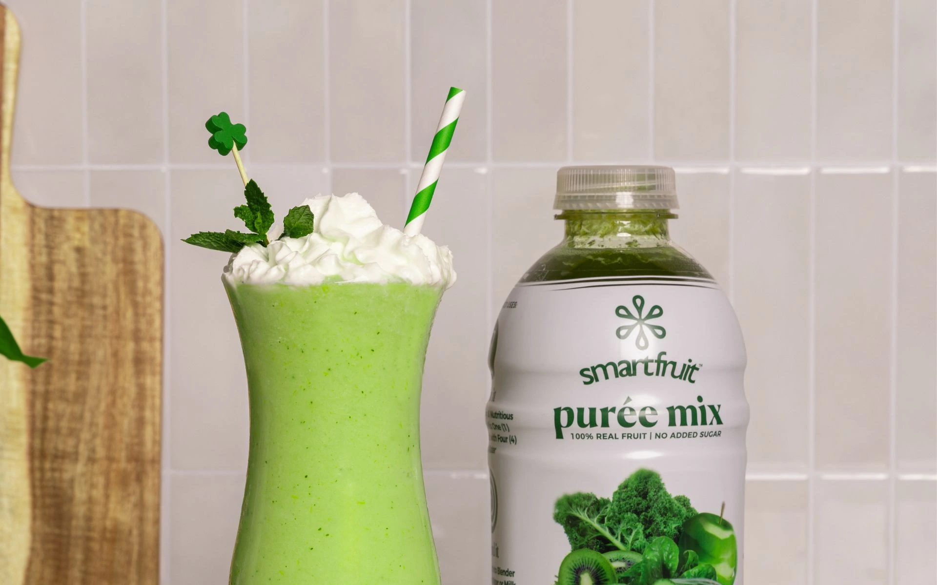 Shamrock shake made with Smartfruit Harvest Greens purée mix, bright green smoothie topped with whipped cream, mint, and a shamrock pick, served next to the purée mix bottle with a striped straw in a clean kitchen setting